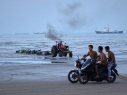 Jóvenes a bordo de motocicletas a lo largo de la orilla mientras un tractor saca equipo del agua y al fondo se pueden ver buques mercantes aparentemente anclados en el estrecho de Ormuz. AP/R. Poudat